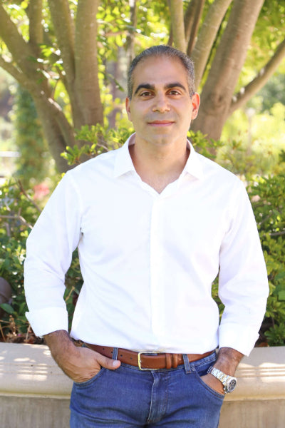 Man wearing a white shirt and blue jeans standing outdoors with greenery in the background